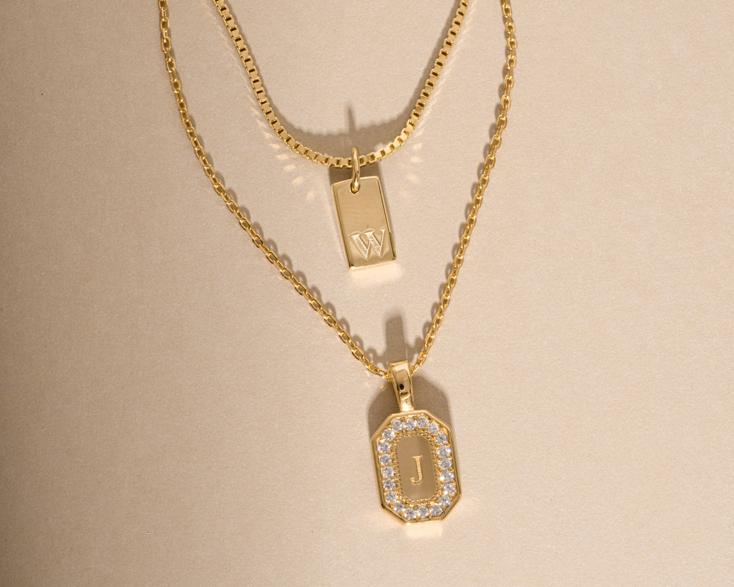 A gold layered necklace set featuring a fine chain and a thicker box chain, both with rectangular initial pendants. The lower pendant is a gold dog tag with a border of small clear stones and a gold letter 'J' in the center, while the upper pendant is a smaller gold block with a matching 'J'.