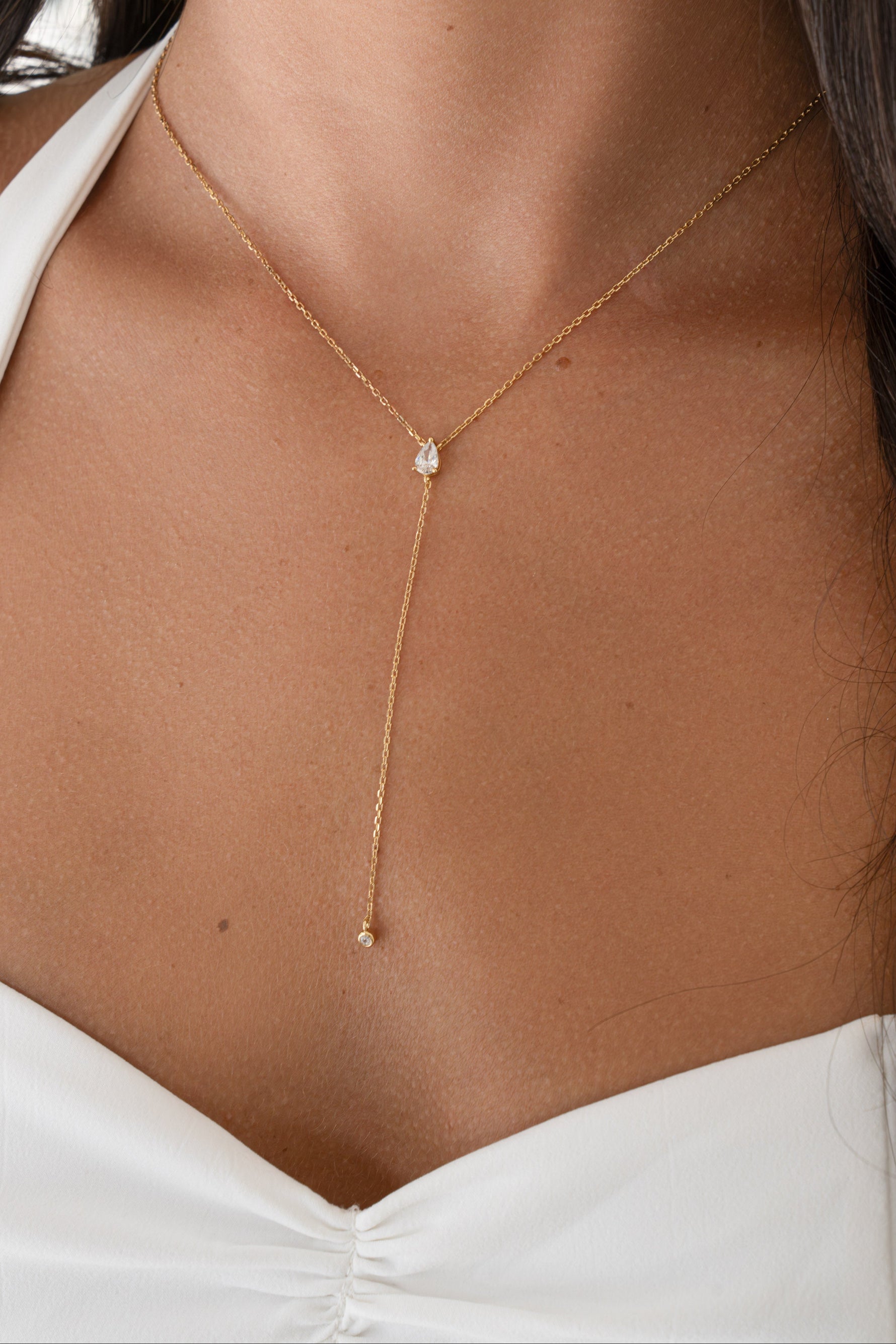 A close-up of a woman wearing a delicate gold Y-necklace with a single small clear stone at the center and a long drop chain ending in a tiny bead, paired with a white halter top.