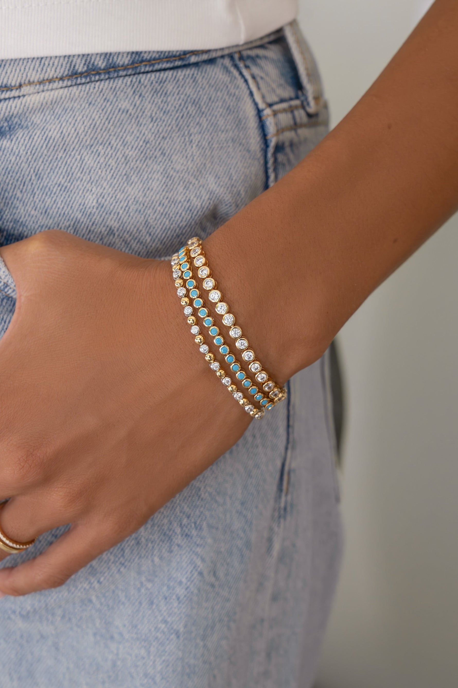 A close-up of a person wearing three delicate beaded bracelets on one wrist, paired with light-wash blue jeans and a white top.
