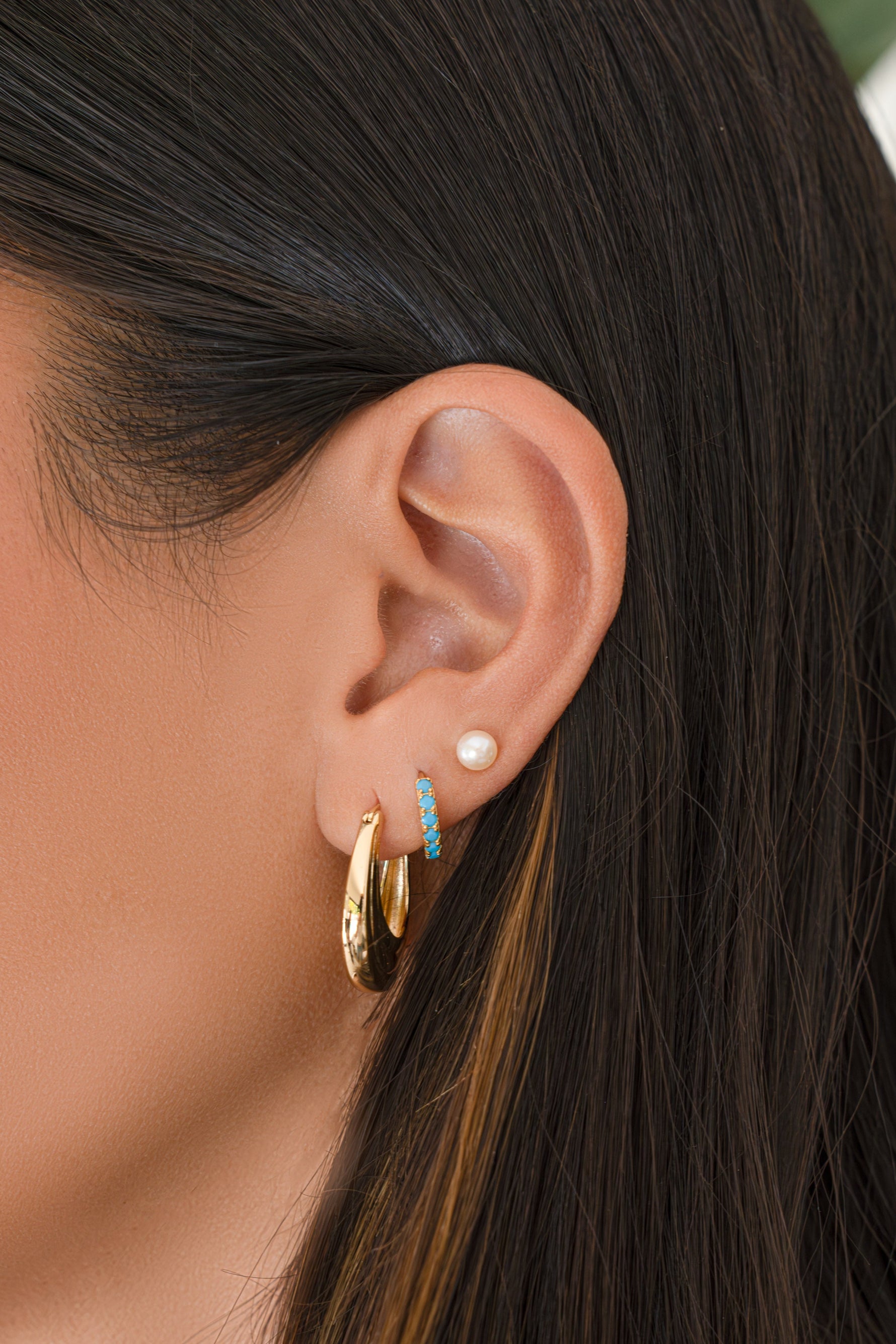 Close-up of a person's ear wearing gold hoop earrings, a small turquoise crescent stud, and a pearl stud, with dark brown hair in the background. 