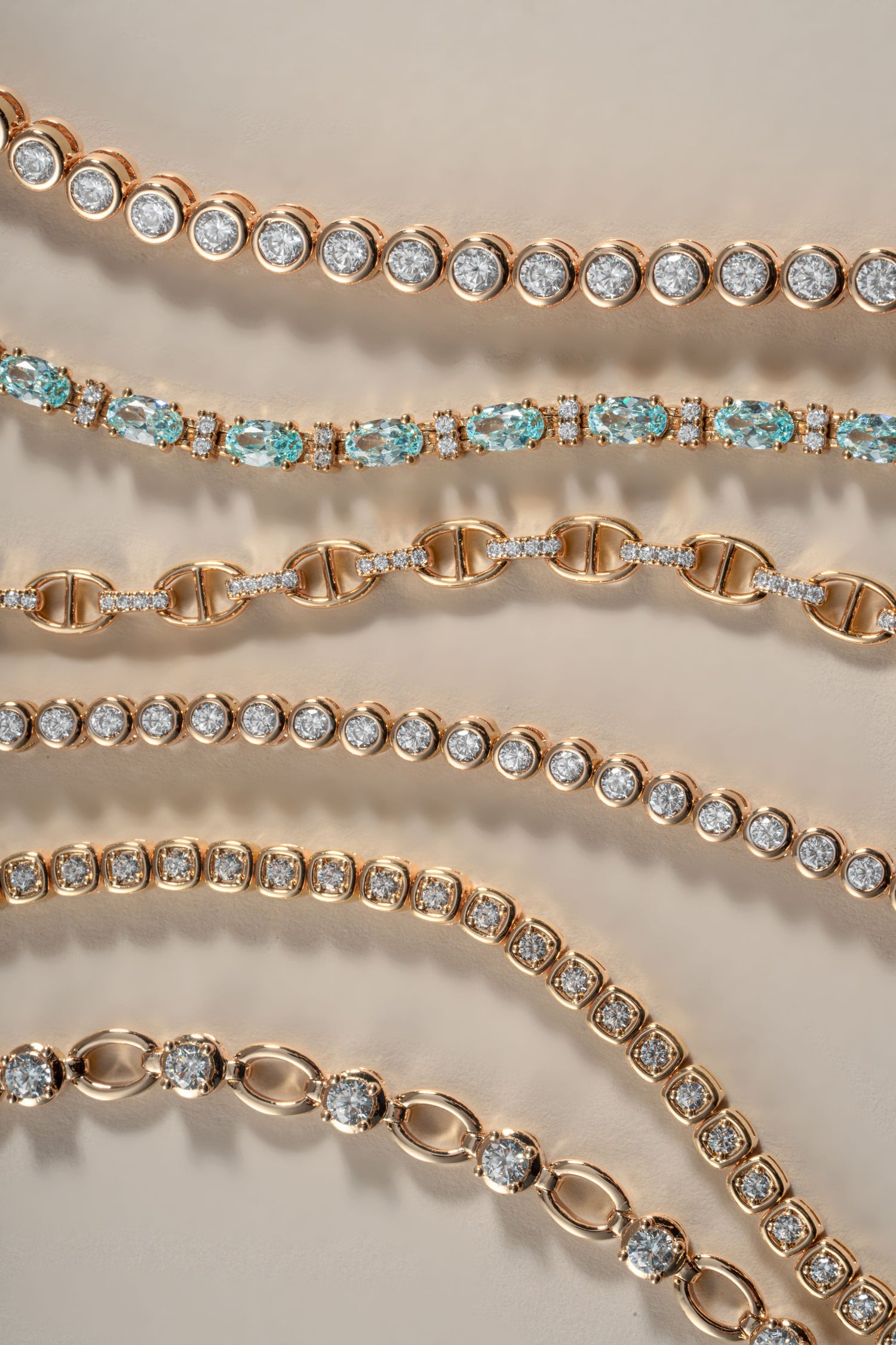 Close-up of multiple rose gold tennis bracelets arranged diagonally on a light beige background. Some bracelets are set with round clear stones, while others feature alternating links with light blue gemstones. 