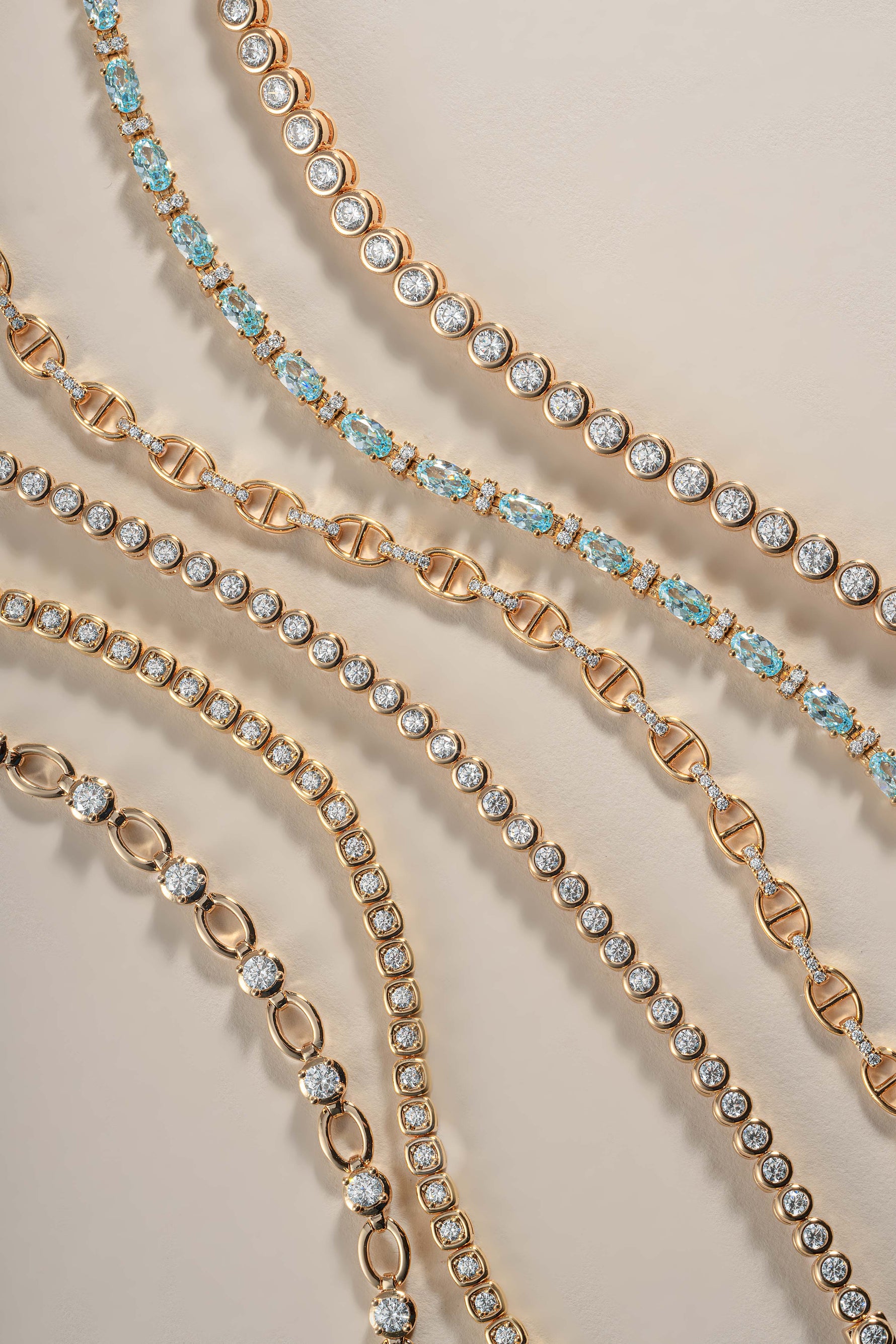 Several gold-toned tennis and link chains with clear stones and light blue gemstone accents arranged diagonally on a warm beige background.