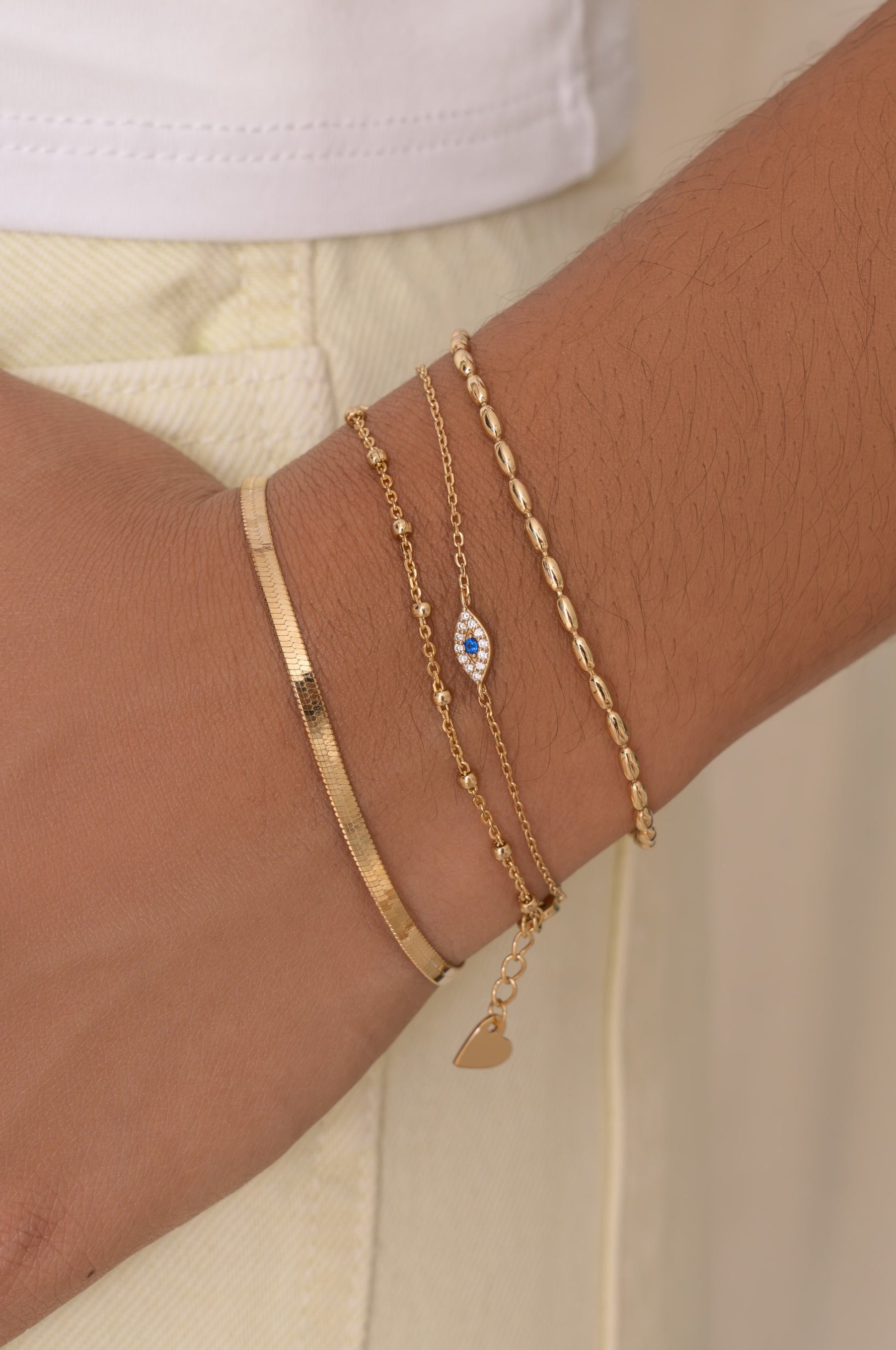 A close-up of a person's wrist wearing multiple delicate gold bracelets, including a flat band bracelet, a chain with an evil eye charm, and a rope-style chain with a small heart charm, paired with a white top and cream pants.