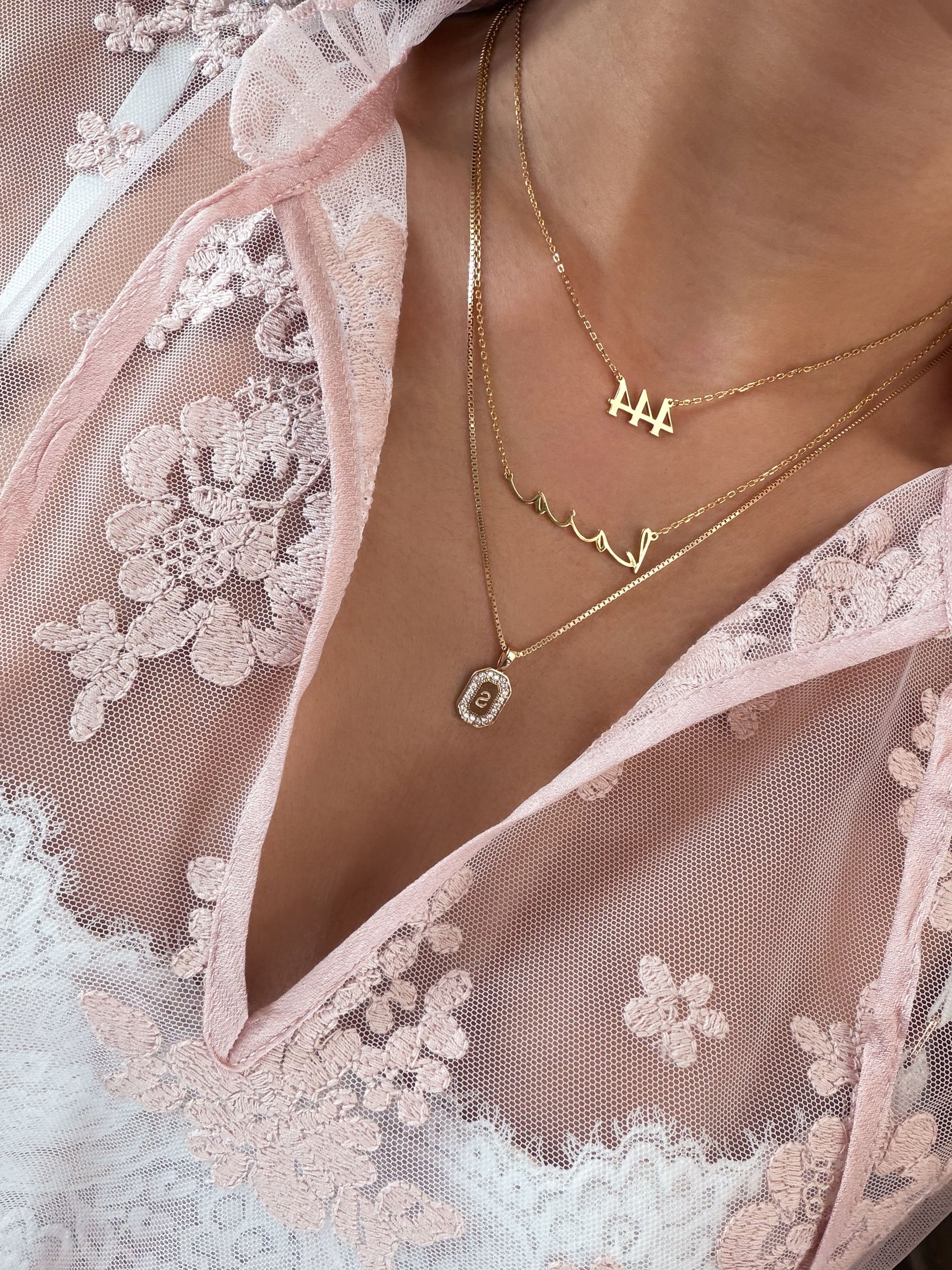 Close-up of a person wearing layered gold necklaces over a sheer light pink lace garment. The necklaces include a small letter H pendant, a script nameplate, and an oval bezel-set stone pendant.