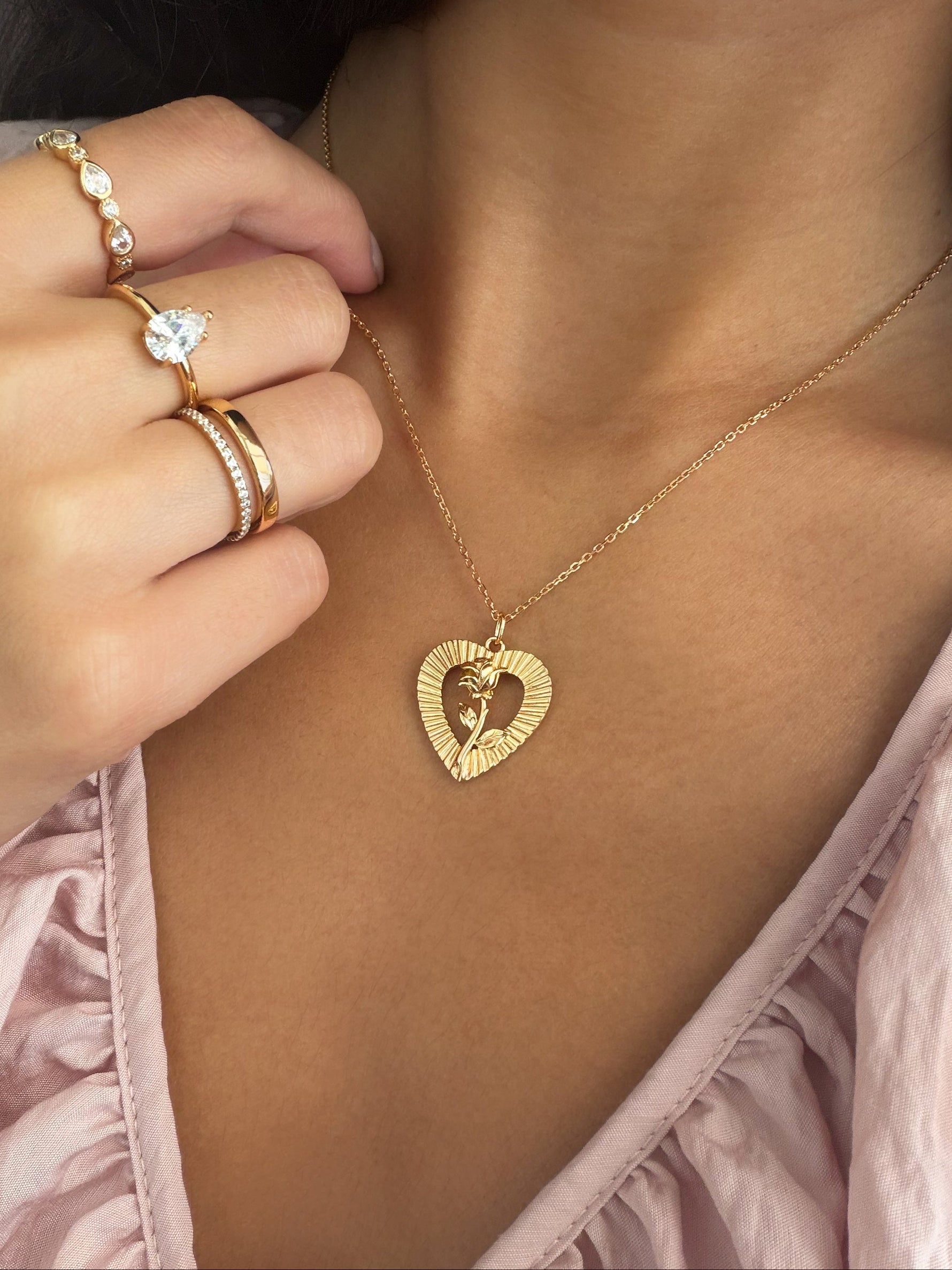 A close-up lifestyle shot of a model wearing a gold heart-shaped pendant necklace with a small cross in the center, styled with gold rings on her fingers.