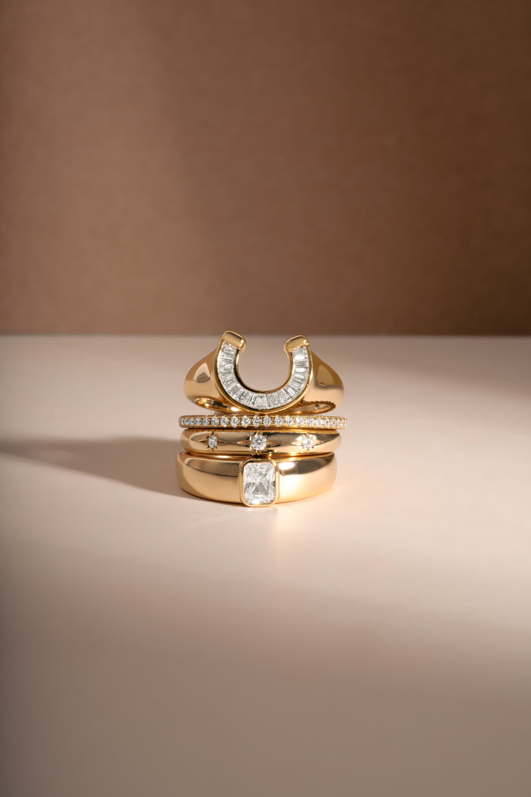 A stack of three gold rings with clear gemstones, including a horseshoe-shaped ring and a ring with a rectangular clear stone, displayed on a beige surface against a brown background.