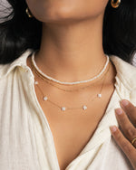 Model wearing three gold necklaces featuring a pearl beaded necklace, a dainty gold beaded necklace, and a diamond flower charm necklace. 
