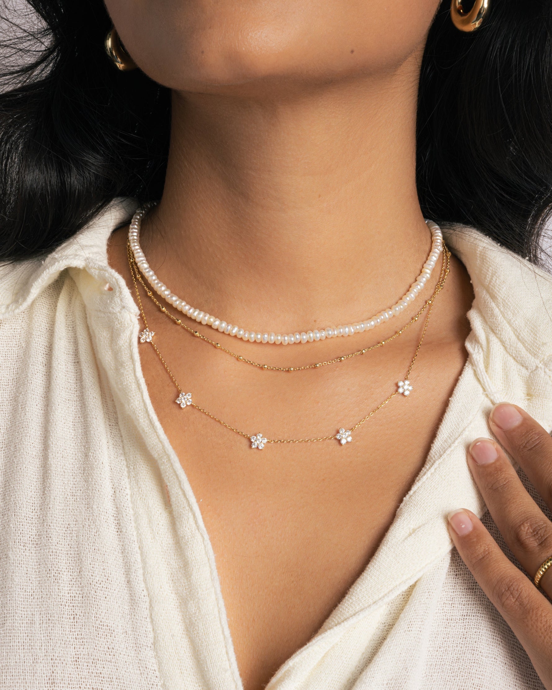 Model wearing three gold necklaces featuring a pearl beaded necklace, a dainty gold beaded necklace, and a diamond flower charm necklace. 
