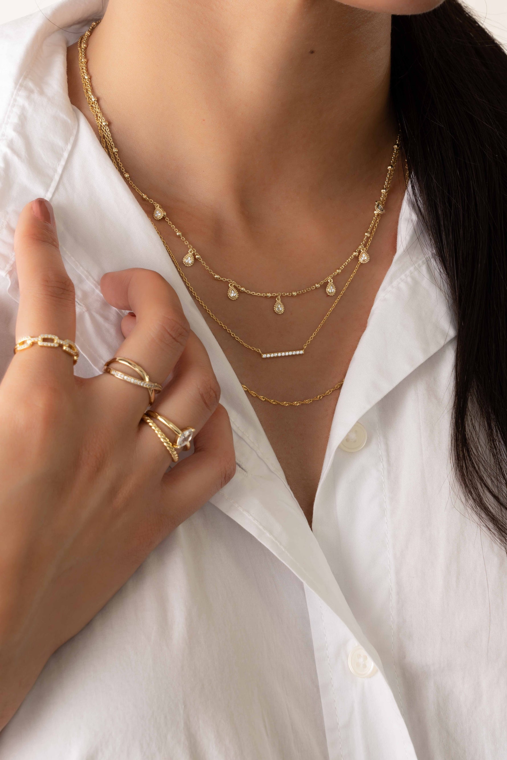 Model wearing dainty tear drop charm necklace stacked with a dainty bar necklace and a thin twisted chain necklace with trendy gold stacking rings. 
