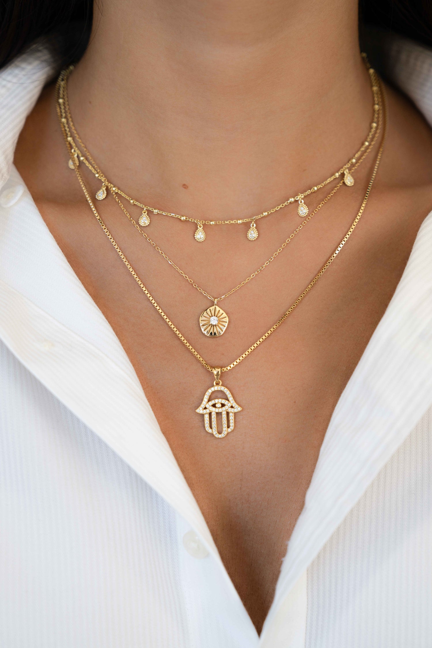 Model wearing three gold necklaces including dangling tear drop charm necklace, a sunburst medallion necklace, and a Hamsa pendant necklace. 