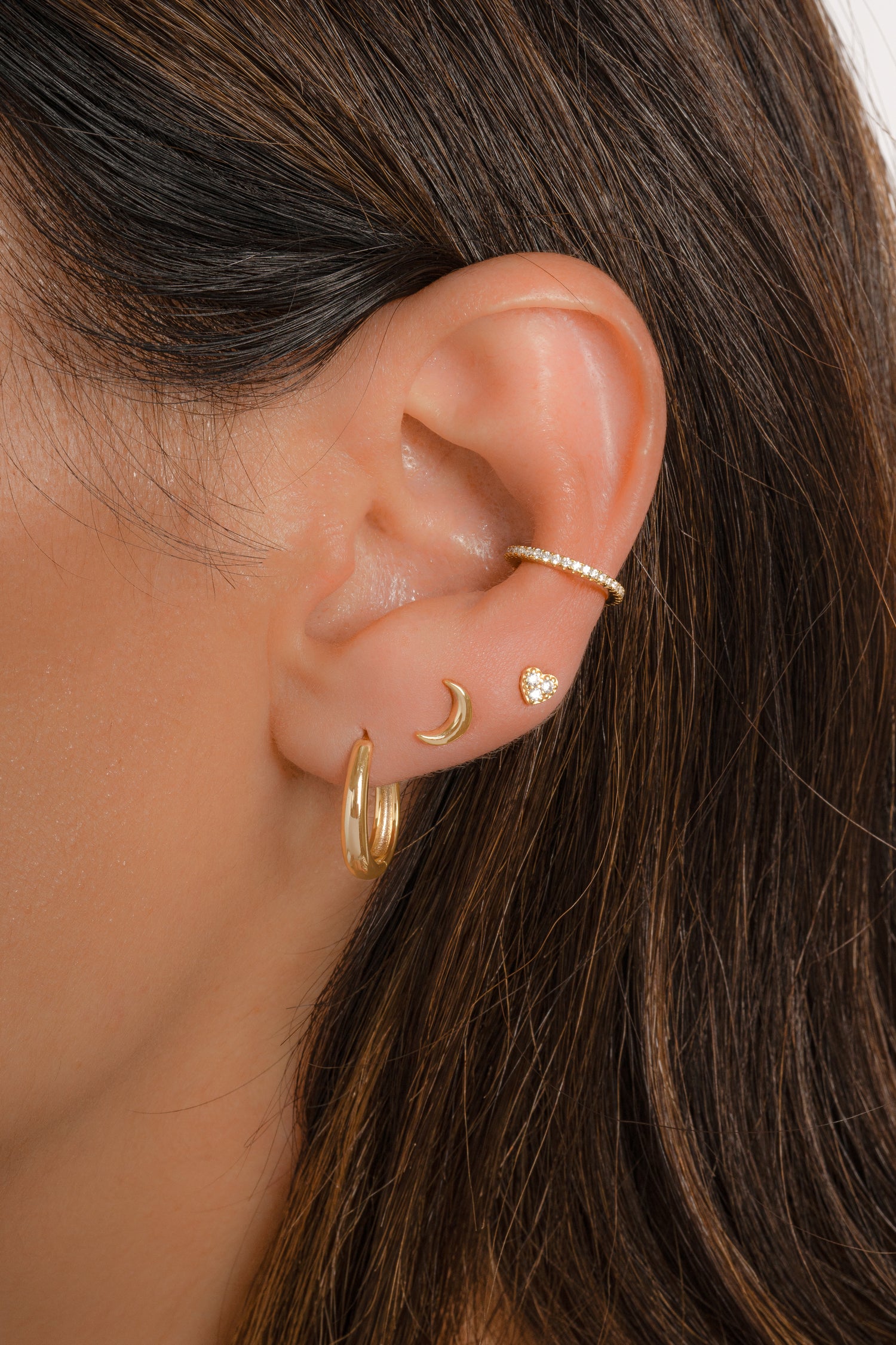 Model wearing dainty gold earring stack with feminine flair, featuring a cz ear cuff, a tiny heart stud, a plain moon stud, and a minimalist oval hoop earring. 