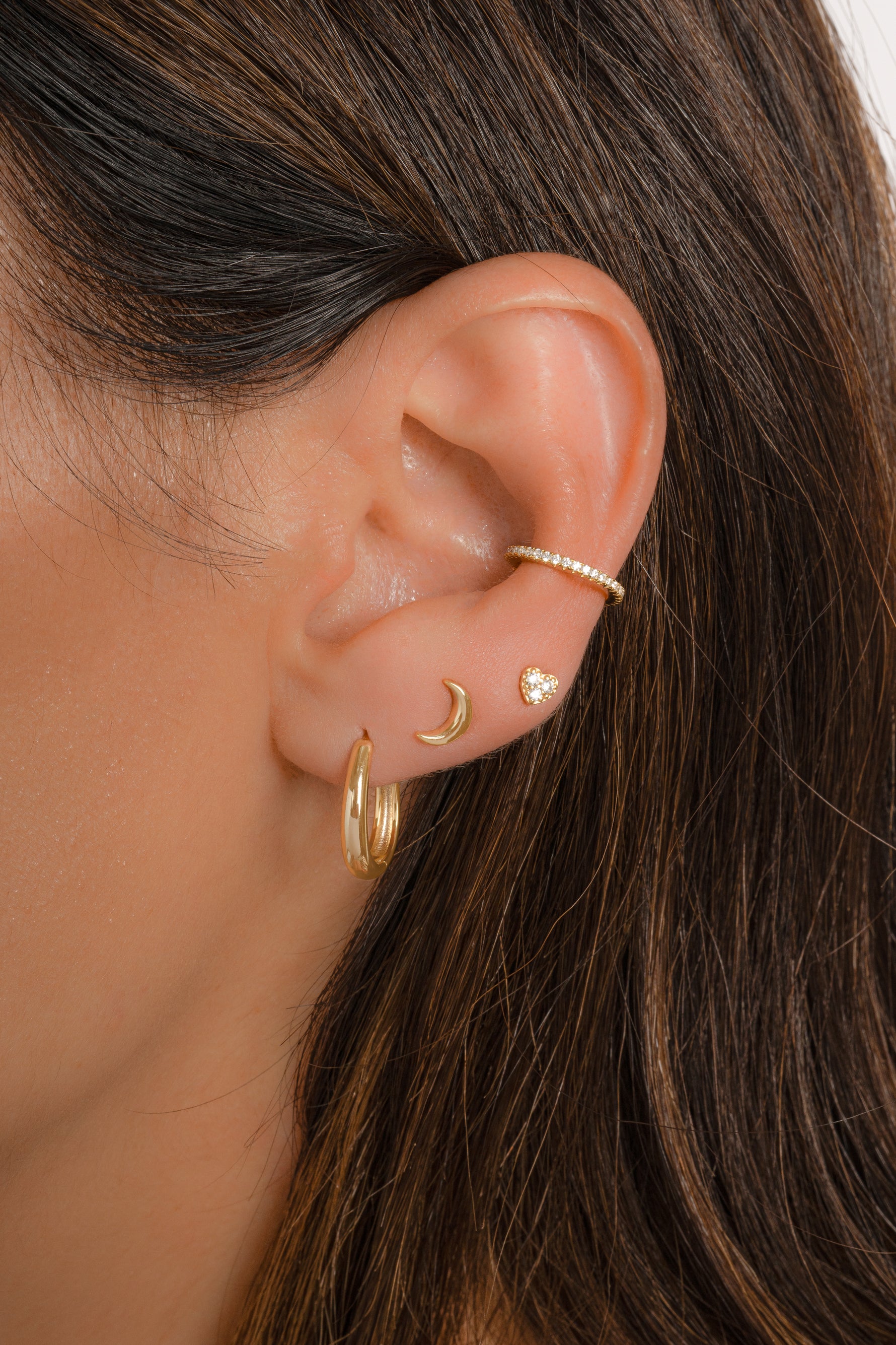 Model wearing dainty gold earring stack with feminine flair, featuring a cz ear cuff, a tiny heart stud, a plain moon stud, and a minimalist oval hoop earring. 