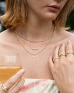 Model wearing three gold layered necklaces including a rice beaded necklace, a gradient bar necklace, and a beaded tear drop necklace. 