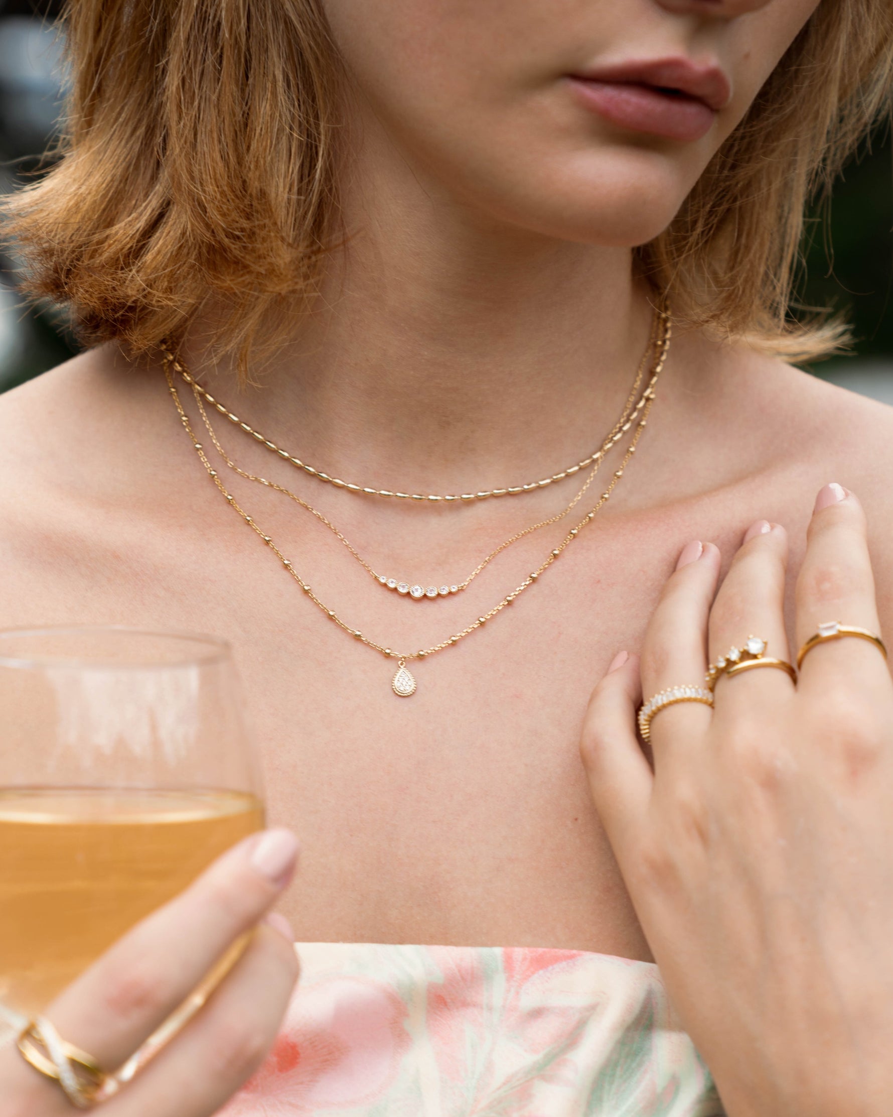 Model wearing three gold layered necklaces including a rice beaded necklace, a gradient bar necklace, and a beaded tear drop necklace. 