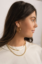 Model wearing sleek minimalist gold jewelry including chunky tube hoops, a gold herringbone chain necklace and a curb link chain necklace. 