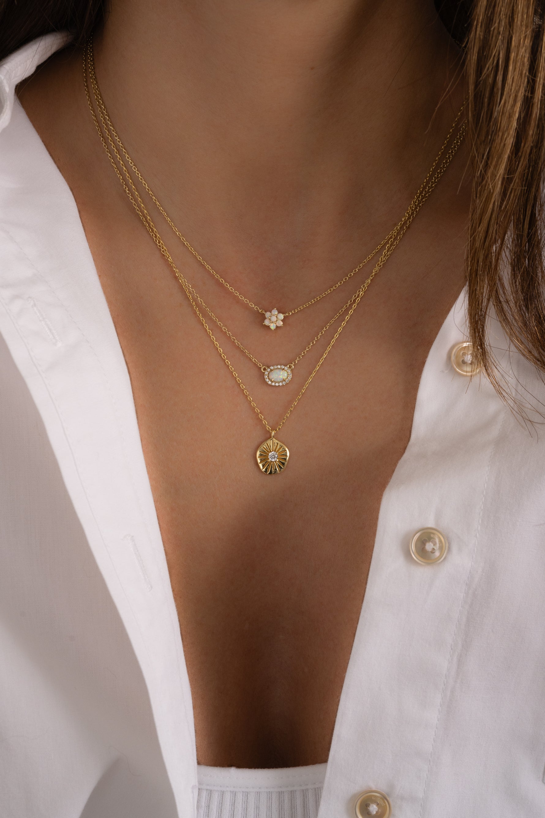Model wearing trendy gold delicate layered pendant necklaces featuring an opal flower necklace, an opal halo necklace, and a sunburst medallion necklace. 