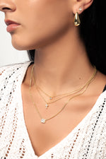 Model wearing trendy and chic gold earrings and necklaces in a stack, featuring a tiny bezel necklace, a cross pendant necklace, and a baguette pendant necklace. 