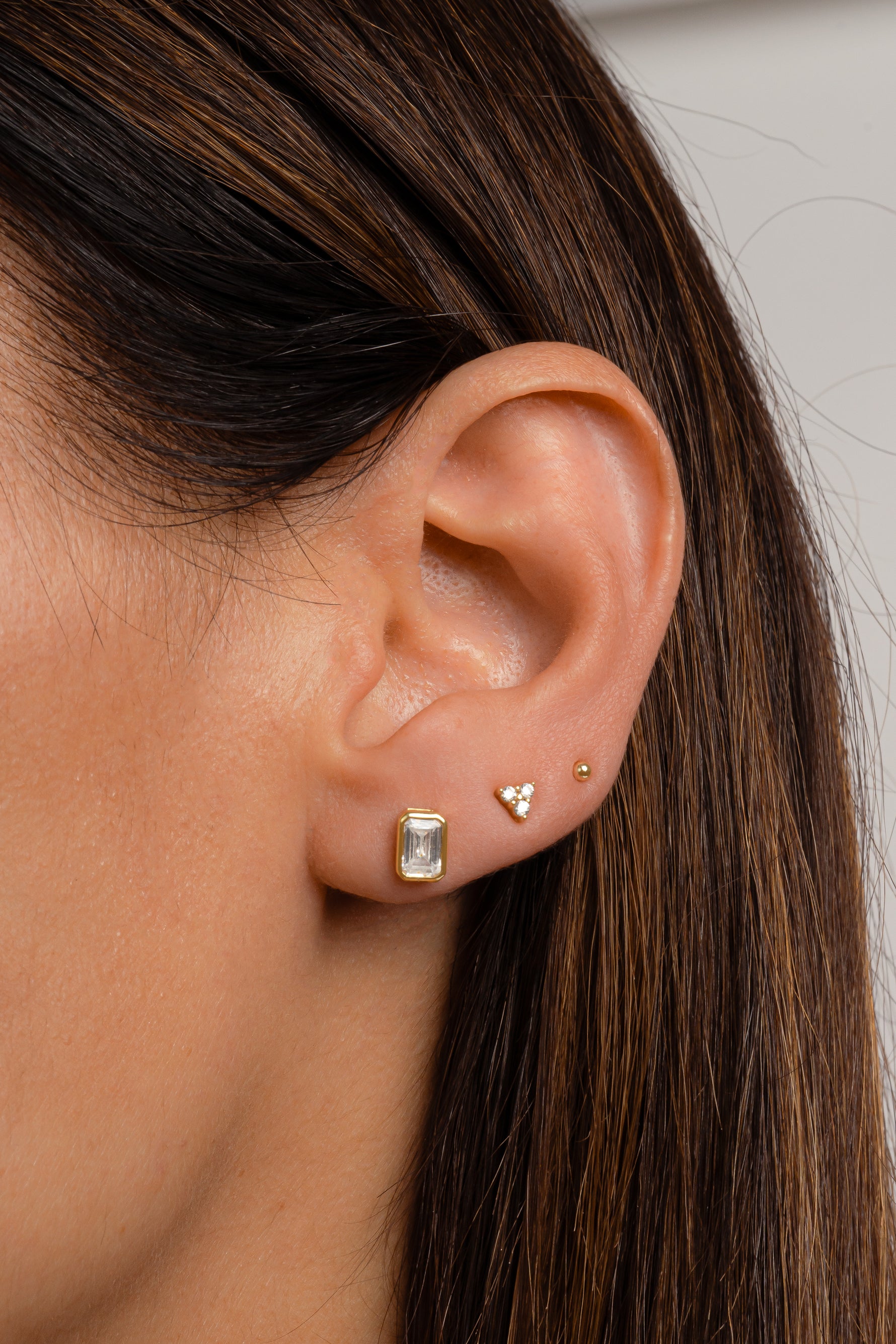 Model wearing gold emerald cut stud earrings paired with 3 stone tiny stud earrings and gold ball stud earrings.