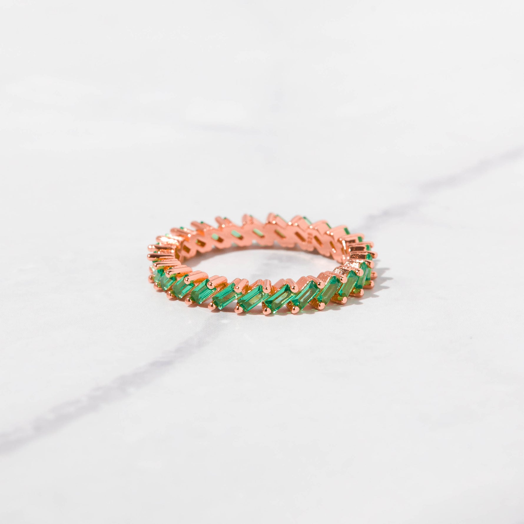 Rose gold emerald slanted eternity ring on marble background. 
