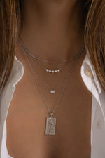 Model wearing layered sterling silver chain necklaces for a chic and trendy look featuring a thin chain necklace, a gradient bezel necklace, a emerald-cut bezel pendant, and a rose rectangle medallion pendant necklace. 