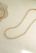A delicate gold tennis necklace with small clear round stones, arranged in a loop on a light cream marble surface with a gold bangle partially visible in the top left corner.