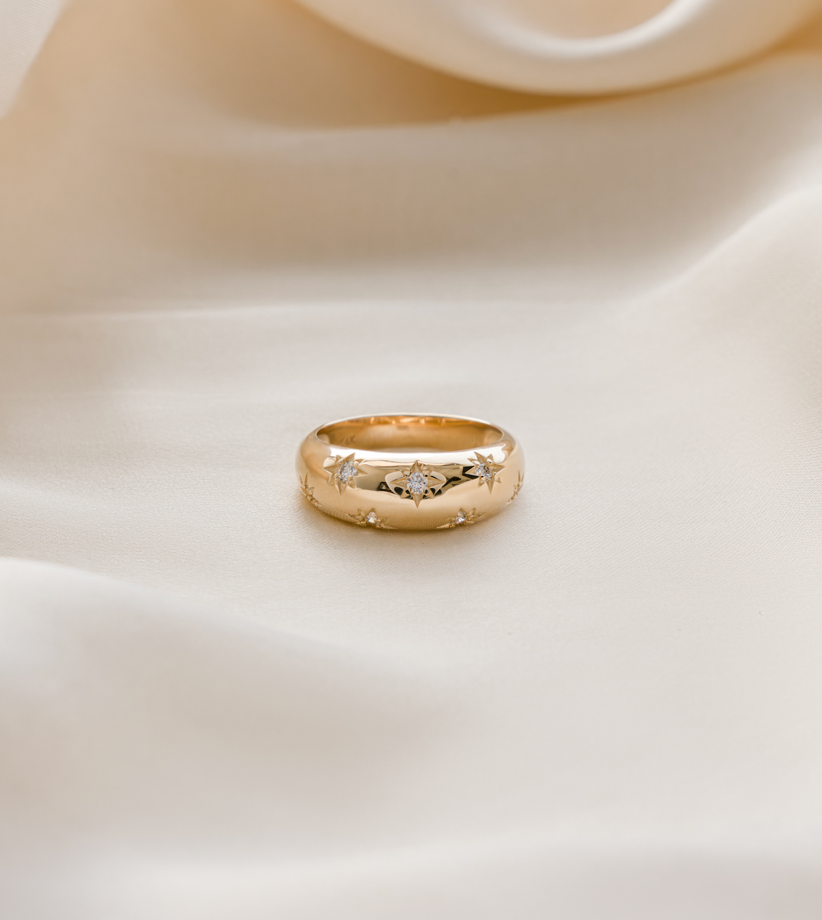 A gold ring with a textured, hammered surface and small clear stones, resting on draped cream-colored satin fabric.