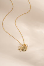 A delicate gold chain necklace featuring a starfish, a round medallion with a palm tree, and a small white pearl, displayed on a soft beige background.