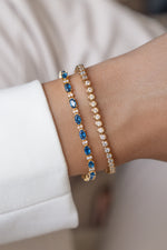 Two gold bracelets featuring a tennis bracelet with oval blue gemstones and alternating round stones, paired with a gold bezel bracelet on wrist.
