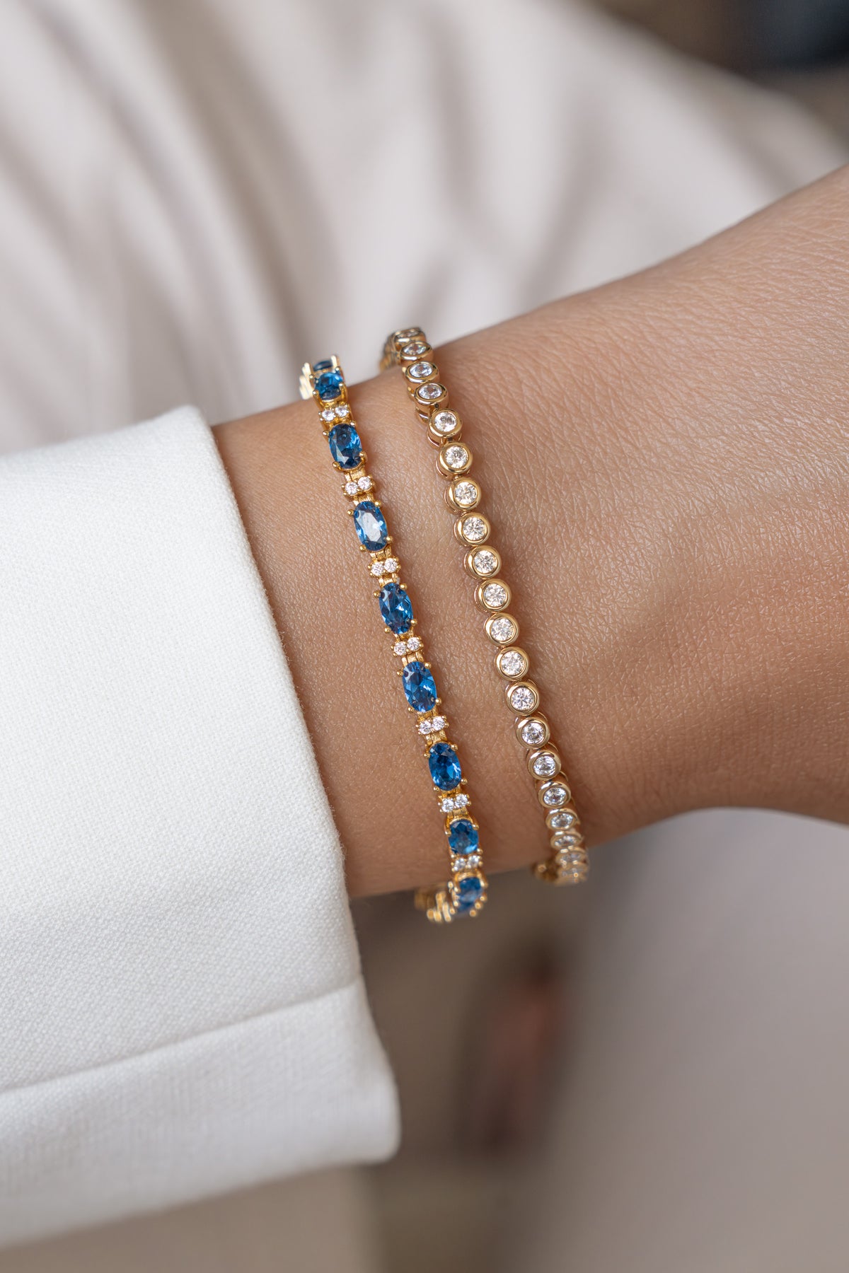 Two gold bracelets featuring a tennis bracelet with oval blue gemstones and alternating round stones, paired with a gold bezel bracelet on wrist.