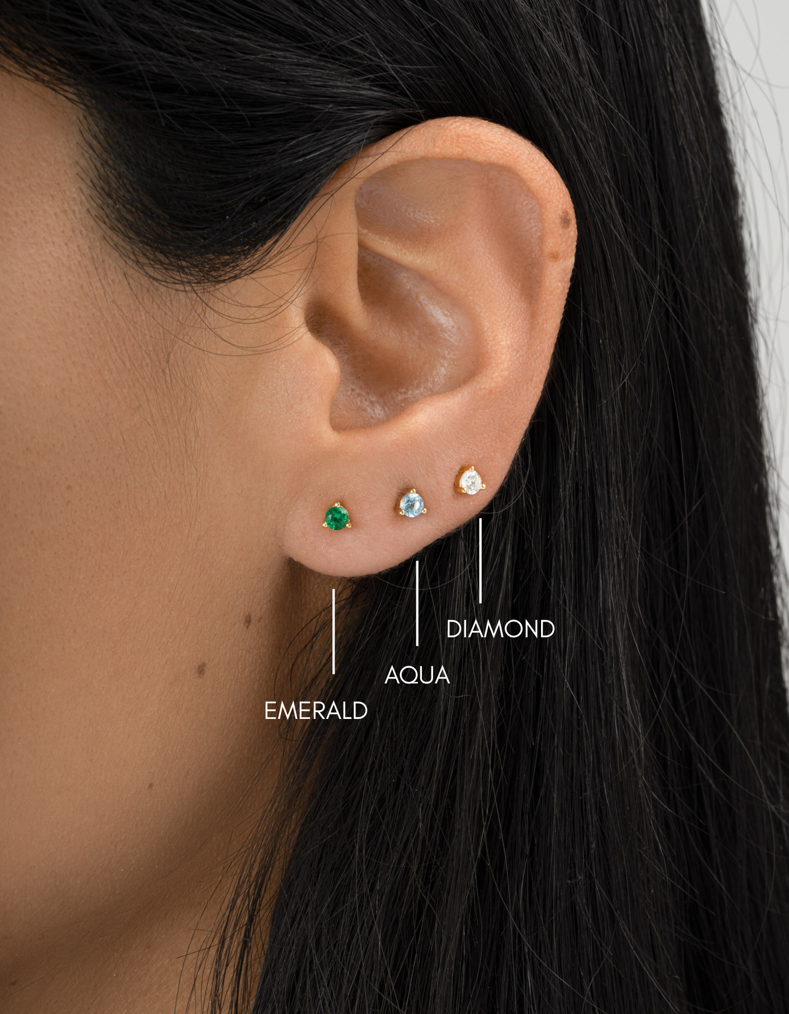 Close-up of an ear wearing three earrings labeled 'Diamond', 'Aqua', and 'Emerald'.