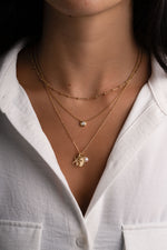 A close-up of a person wearing layered gold necklaces over a white collared shirt. The necklaces include a delicate chain, a small bead pendant, and a longer chain with a starfish charm, a medallion and pearl charm