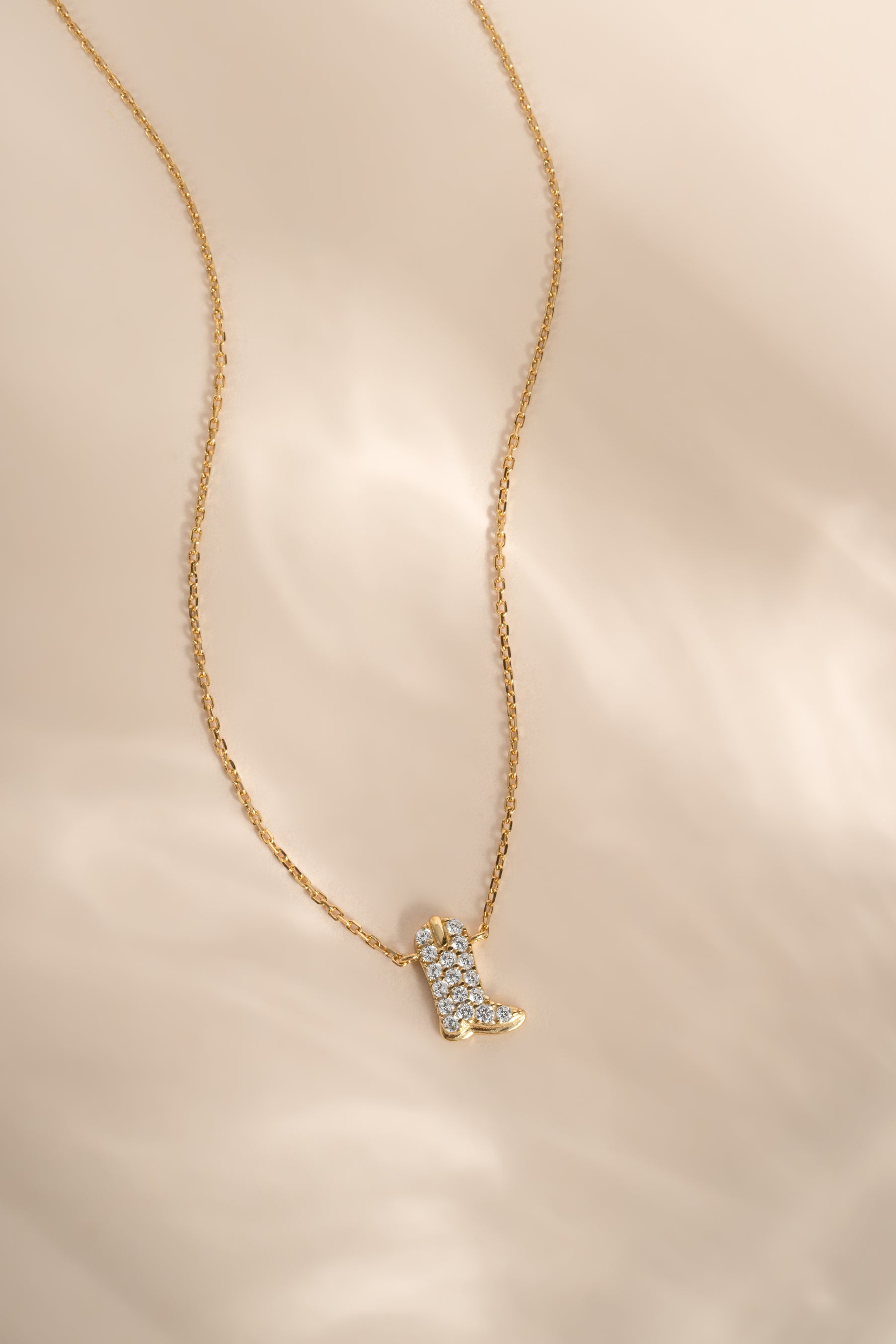 A delicate gold chain necklace featuring a small boot-shaped pendant encrusted with clear stones, displayed on a soft, light beige fabric background.