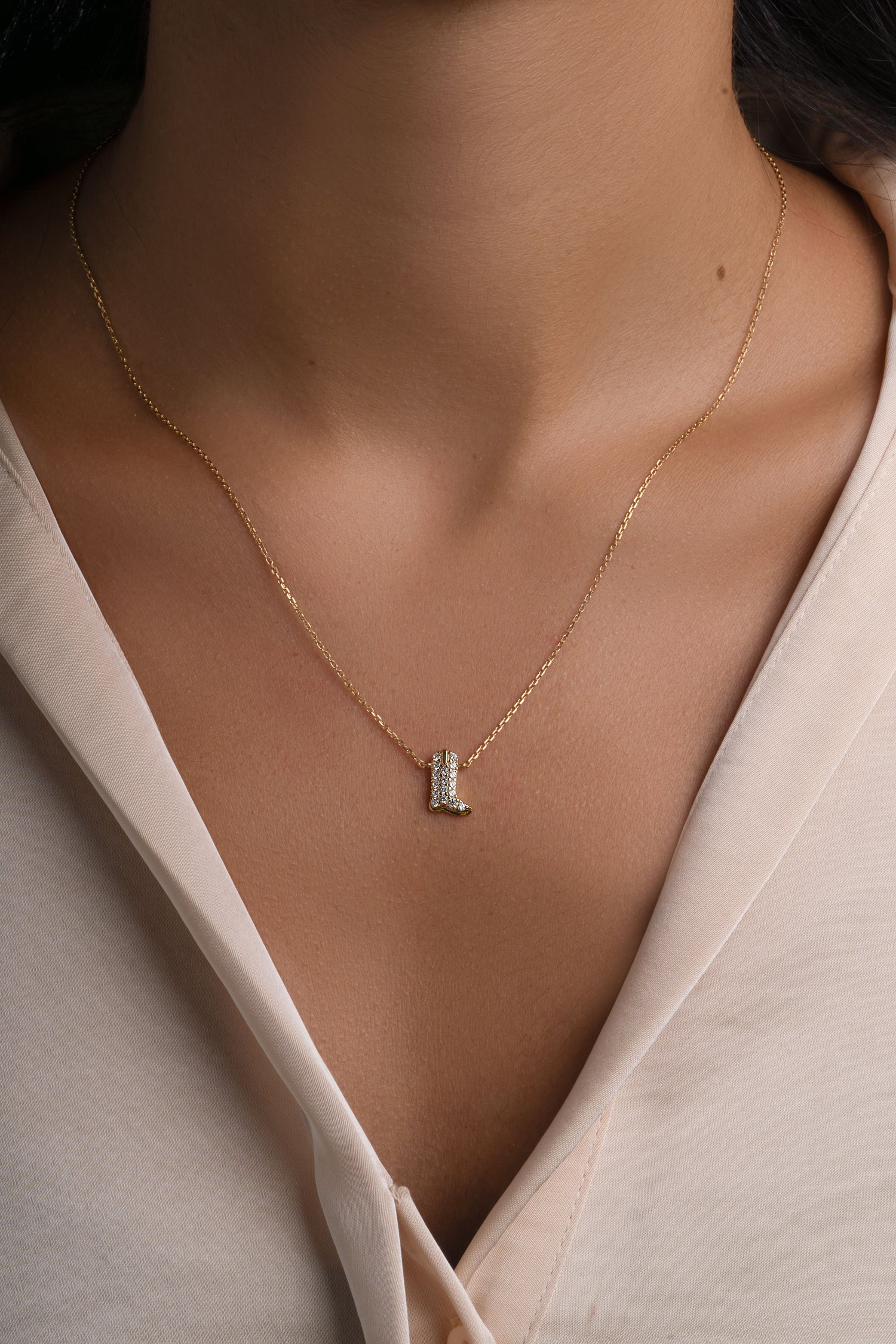 A delicate gold chain necklace with a small cowboy boot pendant featuring clear stones, worn by a model in a cream-colored blouse.