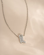 A silver-toned necklace with a cowboy boot pendant paved with clear stones, displayed on a soft beige fabric background.