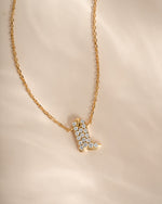 A gold chain necklace with a cowboy boot pendant paved with small clear stones, displayed on a soft beige fabric background.