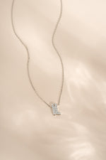 A delicate silver chain necklace featuring a small boot-shaped pendant paved with diamond stones, displayed on a soft, light beige textured fabric background.