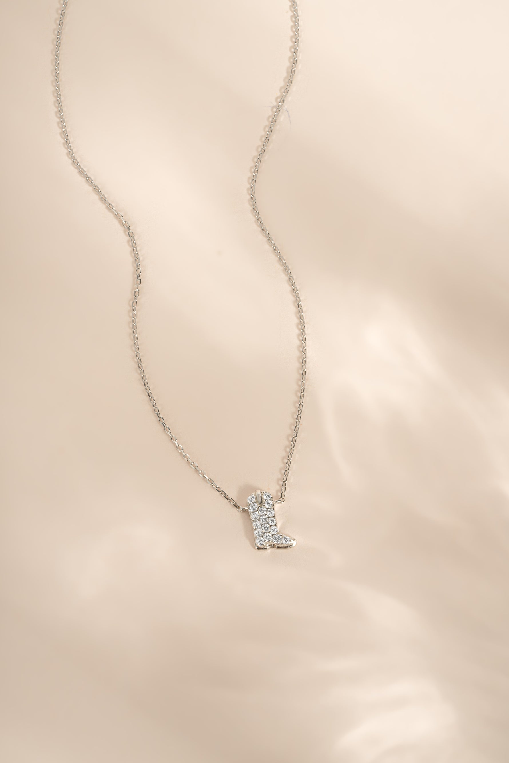 A delicate silver chain necklace featuring a small boot-shaped pendant paved with diamond stones, displayed on a soft, light beige textured fabric background.