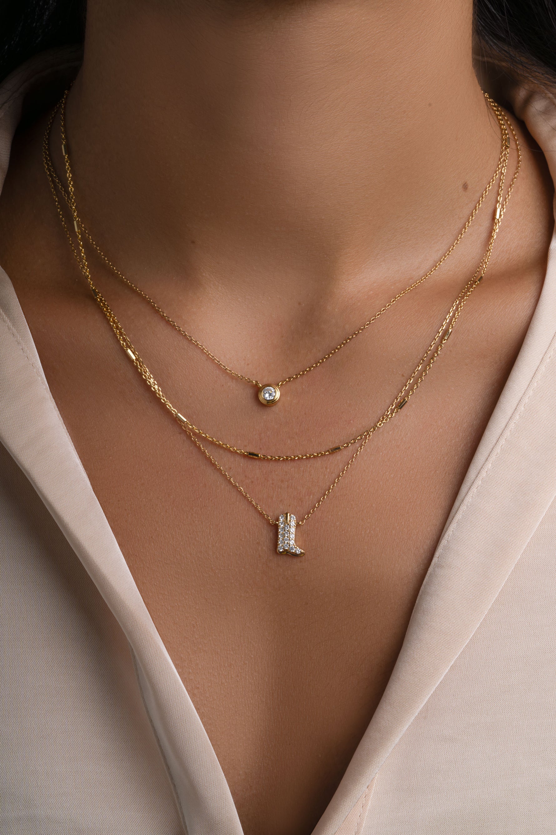 A close-up of a person's neck and upper chest wearing layered gold necklaces, including a small round pendant and a cowboy boot charm pendant with pave diamonds, paired with a cream-colored blouse.