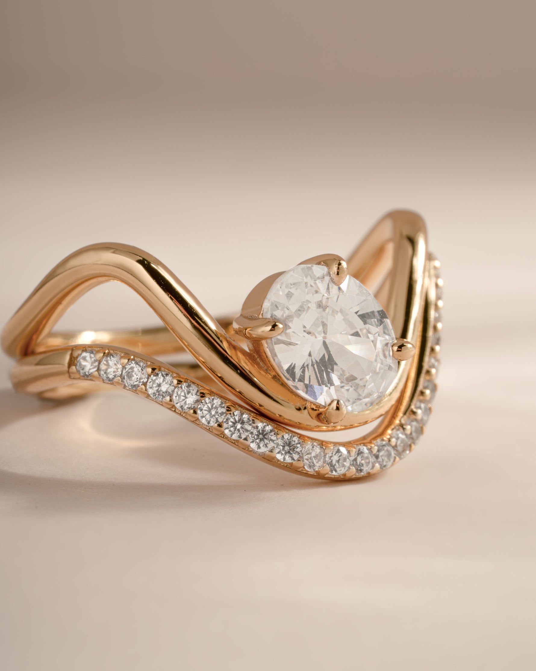 A close-up shot of a rose gold ring featuring a large round brilliant-cut diamond center stone and a curved band adorned with small pavé diamonds, set against a soft beige background.