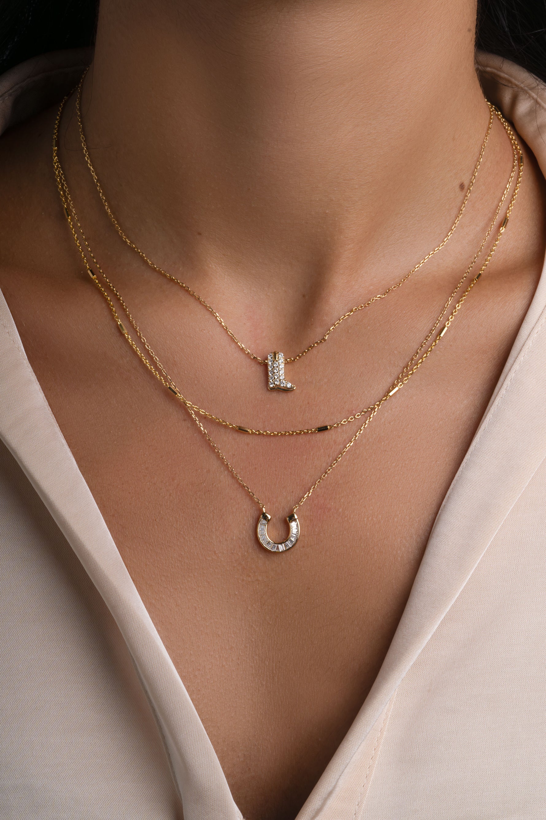 A close-up of a model's neck and chest wearing three layered gold necklaces: a delicate chain necklace with a small cowboy boot pendant, a longer minimalist chain, and a longer chain with a horseshoe pendant set with baguette cubic zirconia stones