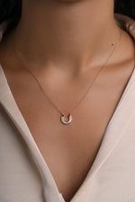 A delicate gold chain necklace with a small horseshoe pendant set with tiny baguette stones, shown worn on a model's neck over a light beige blouse.