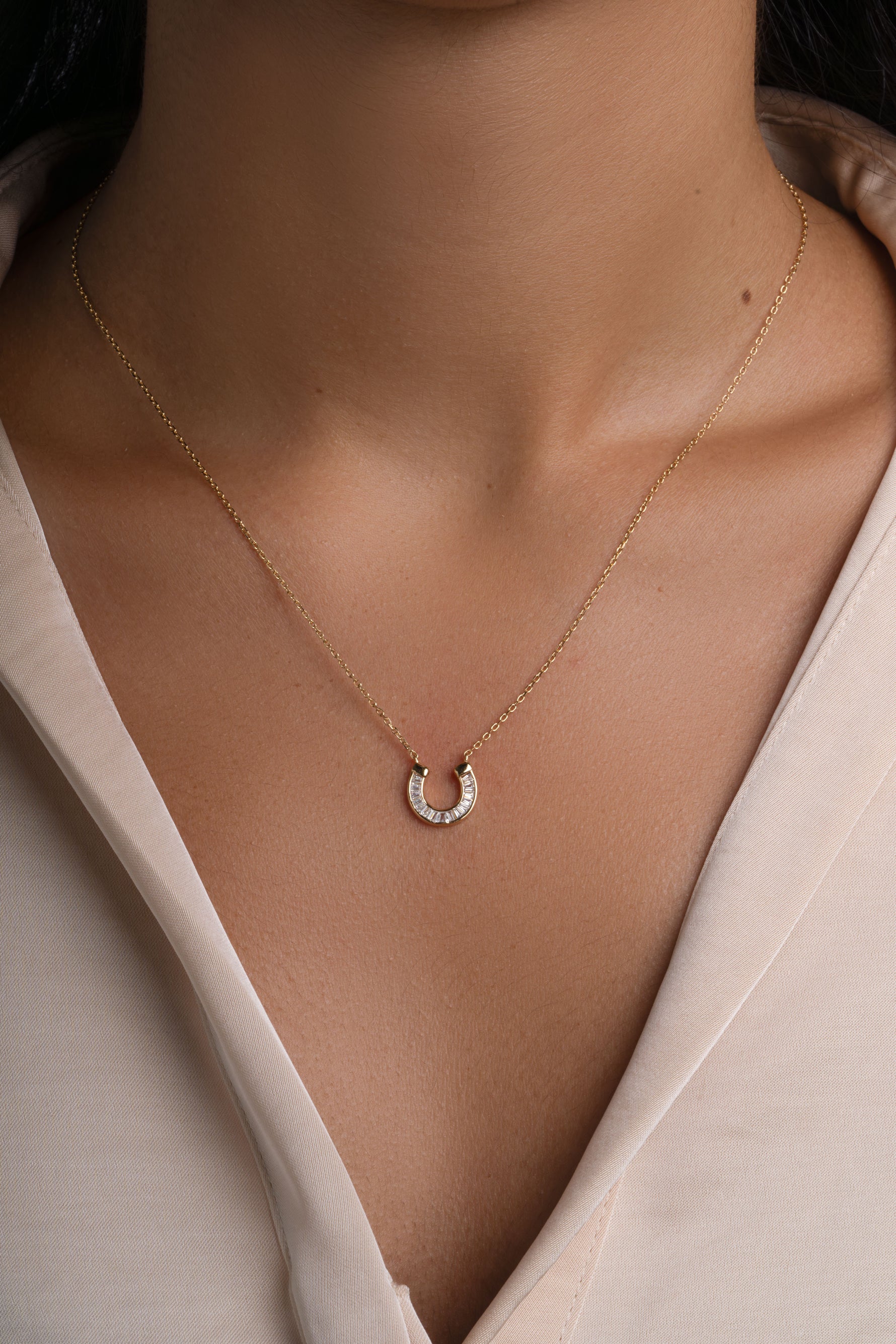 A delicate gold chain necklace with a small horseshoe pendant set with tiny baguette stones, shown worn on a model's neck over a light beige blouse.