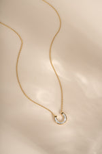 A delicate gold chain necklace with a small horseshoe shaped pendant paved with clear baguette stones, displayed on a soft beige fabric background.