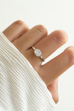 A close-up of a hand wearing a delicate silver ring with a diamond oval stone, styled with a white pleated sleeve against a soft white background.