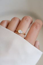 A close-up of a hand wearing a delicate gold ring with an east west oval solitaire cz stone, set against a soft, light beige background with a white sleeve in the foreground.