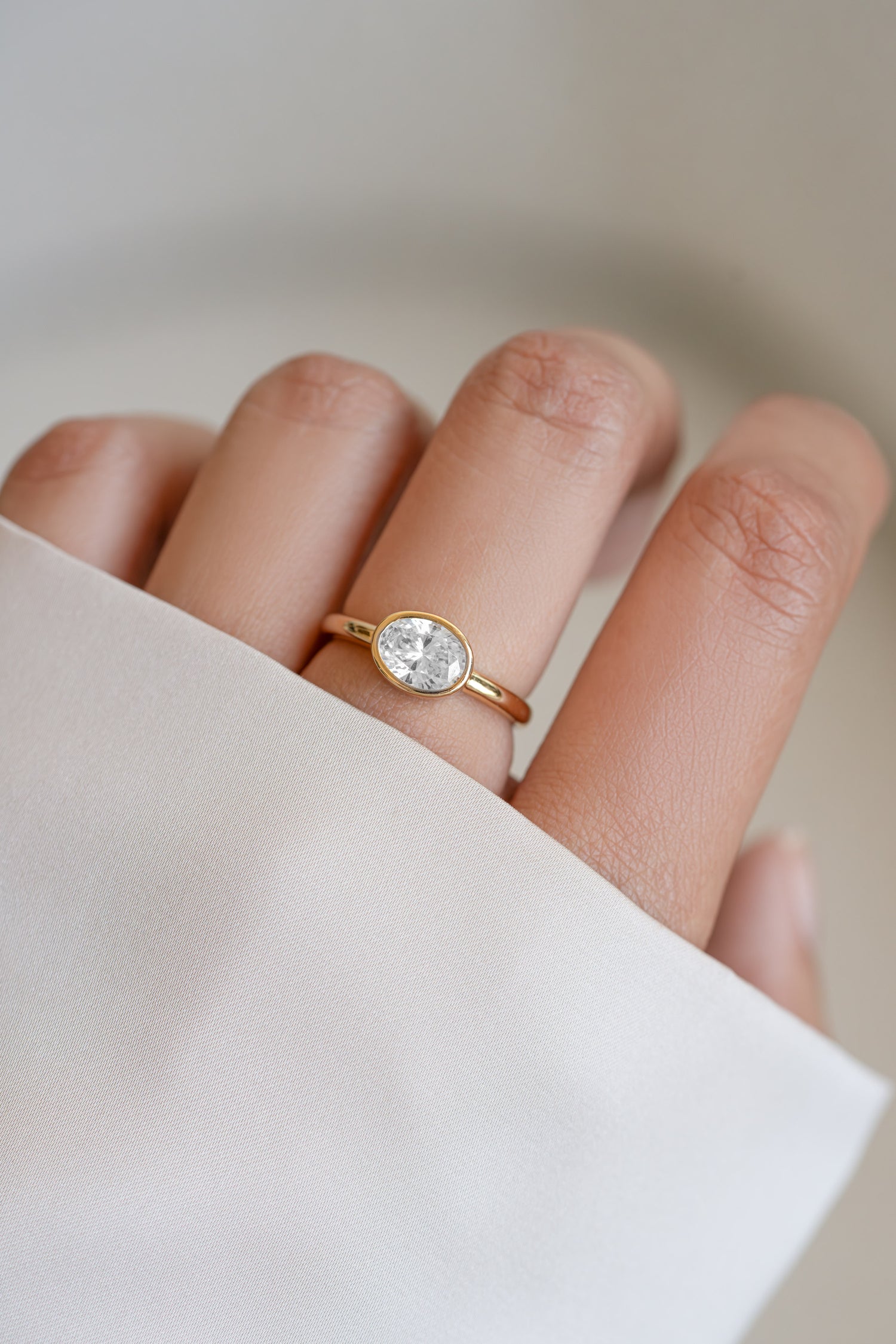 A close-up of a hand wearing a delicate gold ring with an east west oval solitaire cz stone, set against a soft, light beige background with a white sleeve in the foreground.