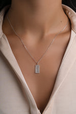 A close-up of a model wearing a delicate silver chain necklace with a rectangular bezel-set clear gemstone pendant with a tropical palm tree engraving detail, styled with a beige satin blouse.