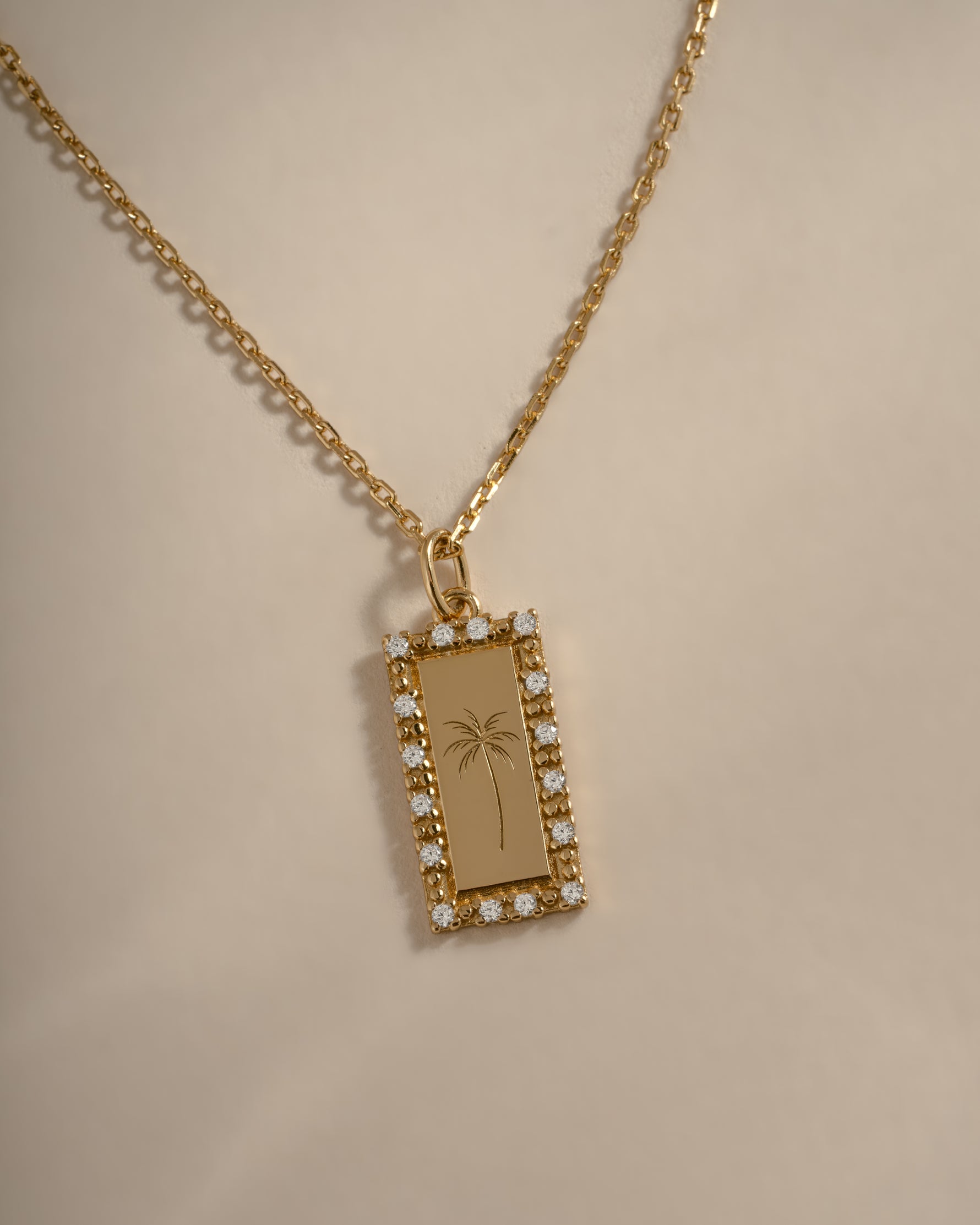 A gold rectangular pendant necklace with a small border of clear stones and an engraved palm tree design, displayed on a beige background.