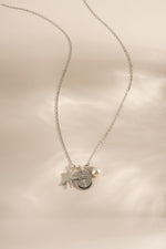A delicate silver chain necklace featuring a circular medallion pendant with a palm tree design, accented by a small pearl bead, and a starfish charm displayed on a soft beige satin background.