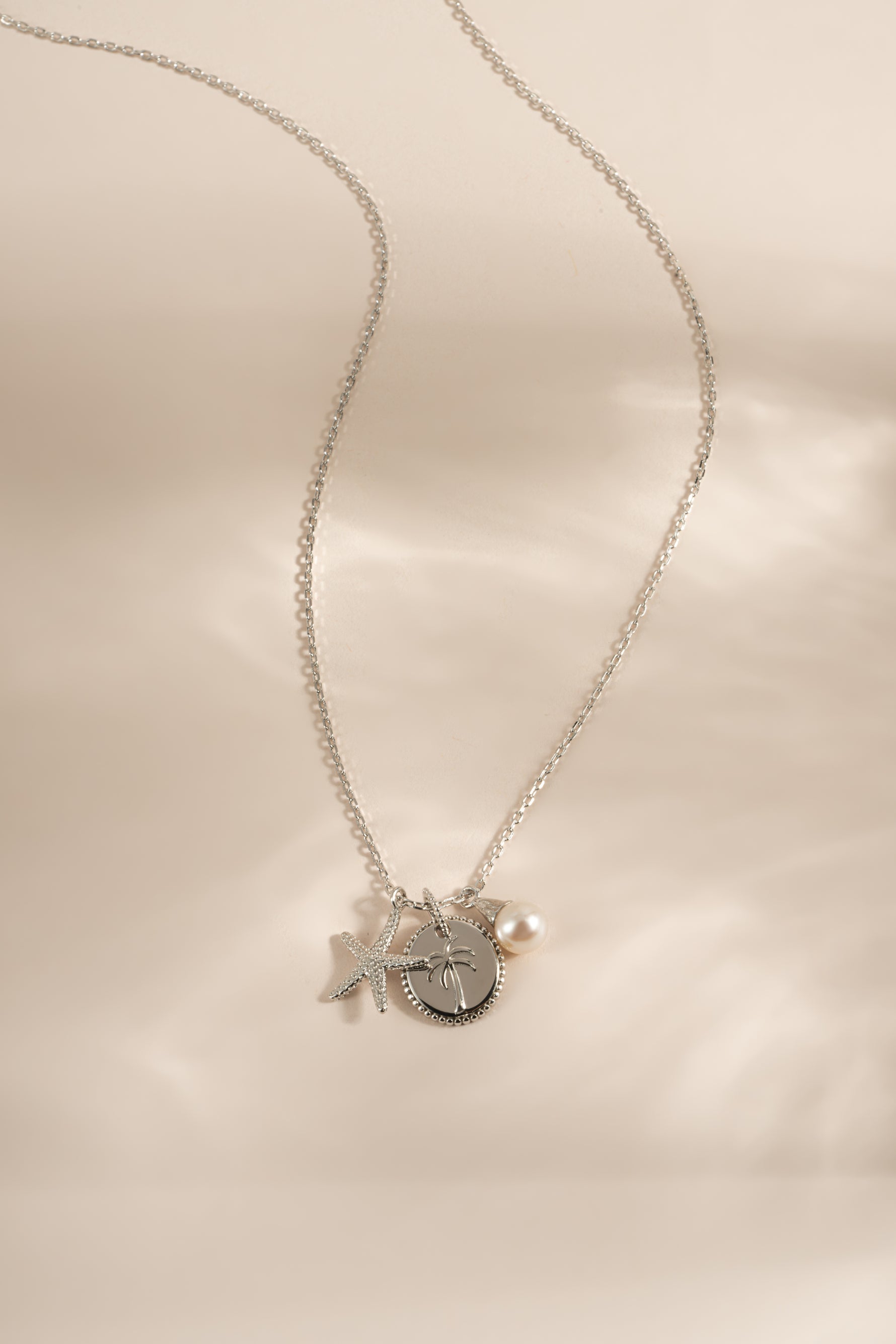 A delicate silver chain necklace featuring a circular medallion pendant with a palm tree design, accented by a small pearl bead, and a starfish charm displayed on a soft beige satin background.